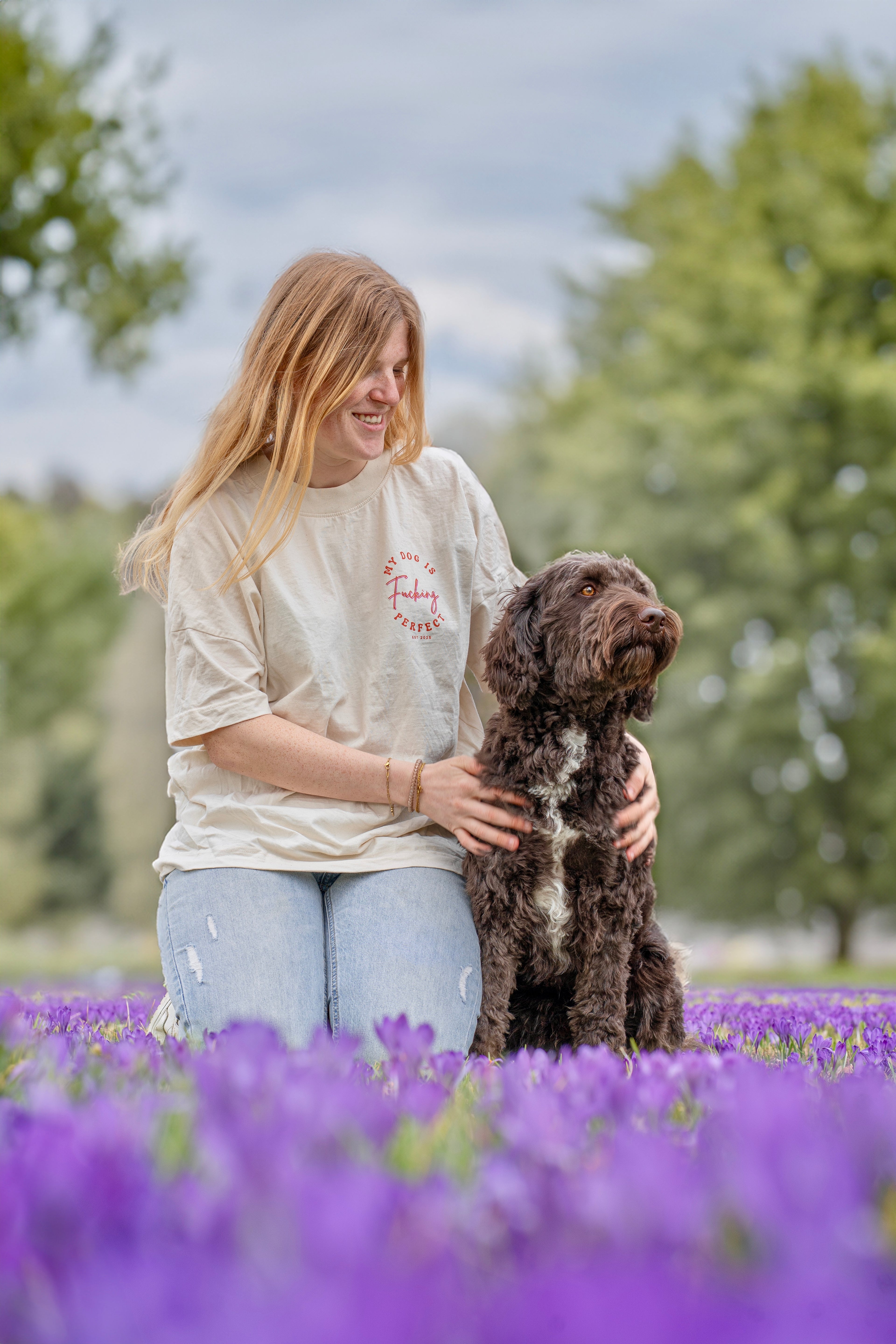 Rothaariges Mädchen spielt mit braun-weißem Labradoodle auf blühender Sommerwiese – fröhliche Hundefotografie